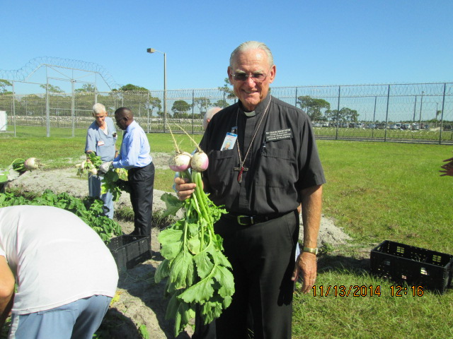 Deacon holding vegetables just harvested by the inmates working in the background.
